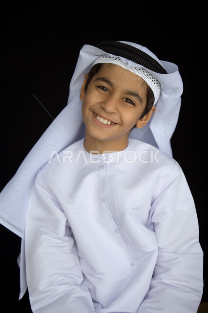 A quarrelsome laugh full of joy, a close-up portrait of a smiling Emirati Gulf Arab boy wearing a white ghutra, a black headband and a traditional kandura, looking at the camera with a tilted head movement, innocent childish features characterized by elegance and handsomeness, a black background