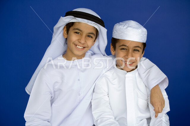 التآلف والمحبة بين الأصدقاء  ، ملامح طفولية بريئة ، بورتريه مقرب  لولدين عربيين  خليجيين إماراتيين  مبتسمين يرتديان الكندورة التقليدية يتعانقان وينظران إلى الكاميرا  ,   قضاء الوقت  مع الأصدقاء بالمرح والتسلية ، خلفية زرقاء .