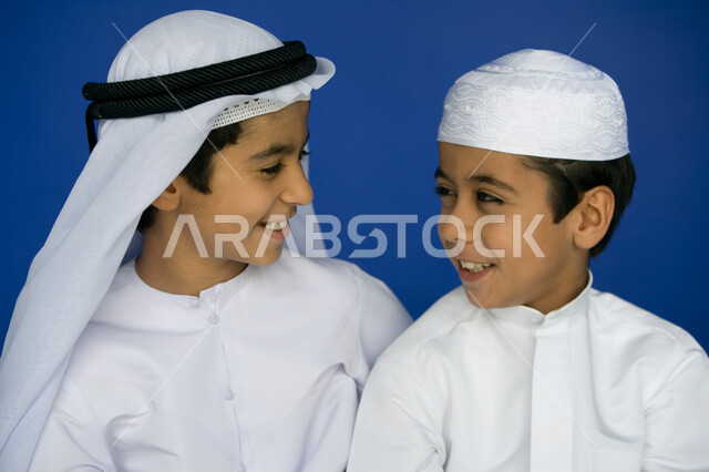 ملامح طفولية بريئة ، بورتريه مقرب  لولدين عربيين  خليجيين إماراتيين  مبتسمين يرتديان الكندورة التقليدية وينظران إلى بعضهما  ,   قضاء الوقت  مع الأصدقاء بالمرح والتسلية ، تعابير الفرح والاستمتاع  ،  خلفية زرقاء .