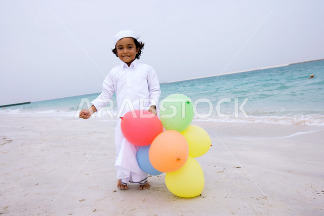 التسلية والترفيه بقضاء أوقات صيفية ممتعة ,  فتى عربي إماراتي خليجي يرتدي الكندورة التقليدية ويقوم باللعب بالبالونات على شاطئ البحر ،الإستمتاع  بفعاليات إجازة الصيف ،  السياحة في دول الخليج العربي ، منتجعات شاطئية في الإمارات ، صورة الجسم بالطول الكامل .