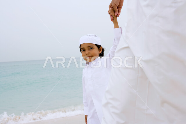Enjoying the activities of the summer vacation, an Arab Emirati Gulf boy holding his father’s hand and wearing the traditional kandora and walking on the seashore, tourism in the Arab Gulf countries, entertainment and entertainment by spending enjoyable summer times, beach resorts in the Emirates