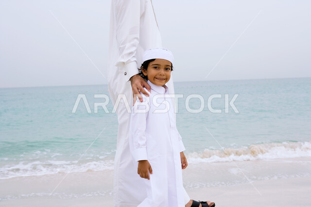 Spending enjoyable summer times, an Arab Emirati Gulf boy accompanied by his father wearing the traditional kandora and walking on the seashore, enjoying summer vacation activities, tourism in the Arab Gulf countries, beach resorts in the Emirates