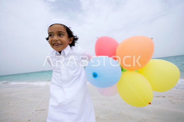 التسالي والمرح بقضاء أوقات صيفية ممتعة ,  فتى عربي إماراتي خليجي يرتدي الكندورة التقليدية ويقوم بالجري  على شاطئ البحر وبيده البالونات ،الإستمتاع  بفعاليات إجازة الصيف ،  السياحة في دول الخليج العربي ، منتجعات شاطئية في الإمارات . 