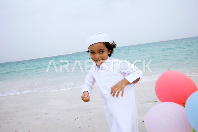 منتجعات شاطئية في الإمارات ،  التسلية والترفيه بقضاء إجازة صيفية ممتعة ,  فتى عربي إماراتي خليجي يرتدي الكندورة التقليدية ويقوم باللعب بالبالونات والجري على شاطئ البحر ،الإستمتاع  بفعاليات إجازة الصيف ،  السياحة في دول الخليج العربي ، صورة الجسم بالطول الكامل .
