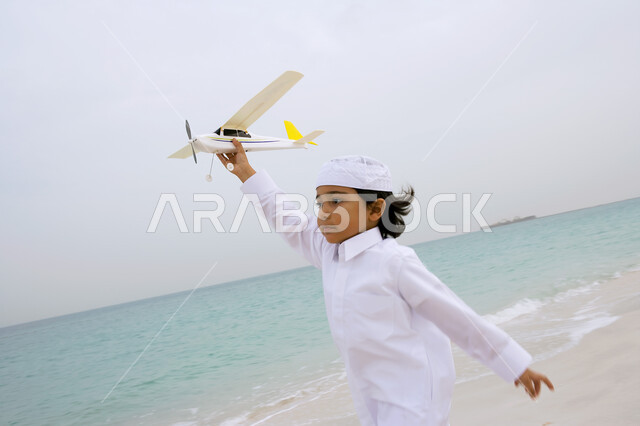 ضحكات مليئة بالسعادة والفرح  ,  فتى عربي إماراتي خليجي يرتدي الكندورة التقليدية ويقوم باللعب بالطائرة على شاطئ البحر ، الإستمتاع  بقضاء إجازة الصيف ،  السياحة في دول الخليج العربي ، منتجعات شاطئية في الإمارات . 