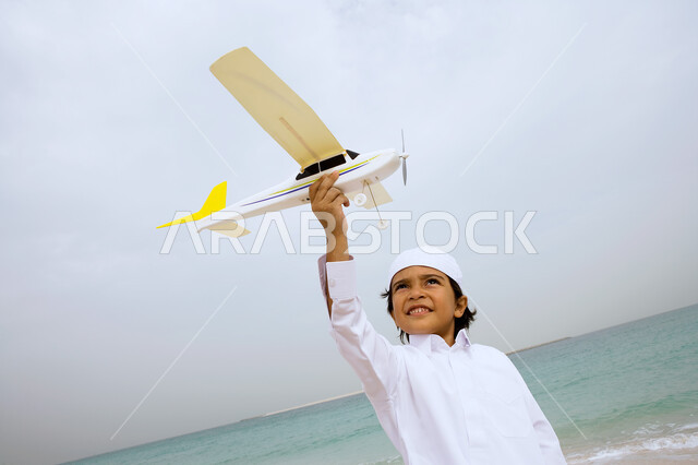 Laughter full of happiness and mixed with joy, an Arab Emirati Gulf boy wearing a traditional kandora and playing with a plane on the seashore, enjoying summer vacation, tourism in the Arab Gulf countries, beach resorts in the Emirates