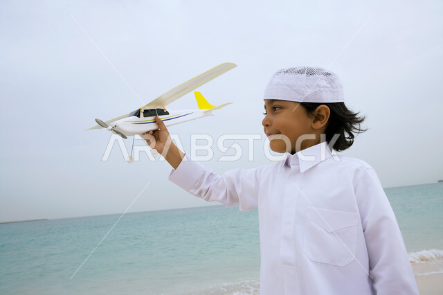 الإستمتاع  بقضاء إجازة الصيف ، ضحكات مليئة بالسعادة والفرح  ,  فتى عربي إماراتي خليجي يرتدي الكندورة التقليدية ويقوم باللعب بالطائرة على شاطئ البحر ،   السياحة في دول الخليج العربي ، منتجعات شاطئية في الإمارات . 