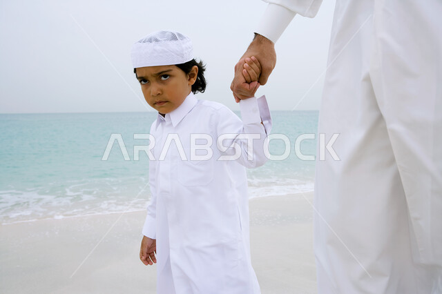 التسلية والترفيه بقضاء أوقات الإجازة الصيفية  ,  فتى عربي إماراتي خليجي يرتدي الكندورة التقليدية يمسك يد والده ويسيران على شاطئ البحر ،الإستمتاع  بقضاء لحظات عائلية ممتعة ،  السياحة في دول الخليج العربي ، منتجعات شاطئية في الإمارات . 