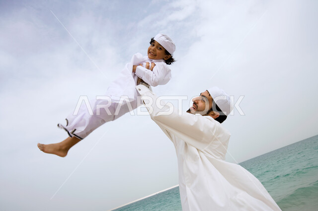 مشاركة الأبناء نشاطاتهم والمرح معهم ,  رجل عربي إماراتي خليجي يرتدي الكندورة التقليدية ويرفع ابنه عاليا في السماء على شاطئ البحر ،قضاء أوقات عائلية ممتعة في الإجازة الصيفية  ،  السياحة في دول الخليج العربي ، منتجعات شاطئية في الإمارات . 
