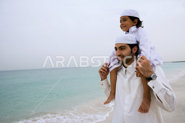 Participating with children in playing and having fun, an Emirati Gulf Arab man wearing a traditional kandora and carrying his son on his shoulders on the seashore, enjoying summer vacation activities, tourism in the Arab Gulf countries, the concept of a happy family and the love of parents for their children