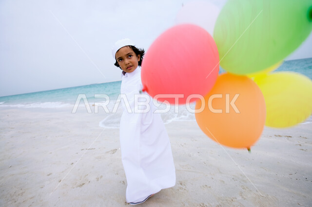 Entertainment and entertainment by spending enjoyable summer times, an Arab Emirati Gulf boy wearing the traditional kandora and playing with balloons on the seashore, enjoying summer vacation activities, tourism in the Arab Gulf countries, beach resorts in the Emirates, full-length body image