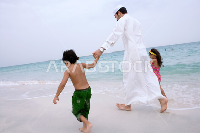 التشارك في اللعب والمرح مع الأبناء   ,  رجل عربي إماراتي خليجي يرتدي الكندورة التقليدية ويمسك بأيدي ولديه  ويركضان معا على شاطئ البحر  الرملي ،الإستمتاع  بفعاليات إجازة الصيف ،  السياحة في دول الخليج العربي ، لحظات عائلية سعيدة .