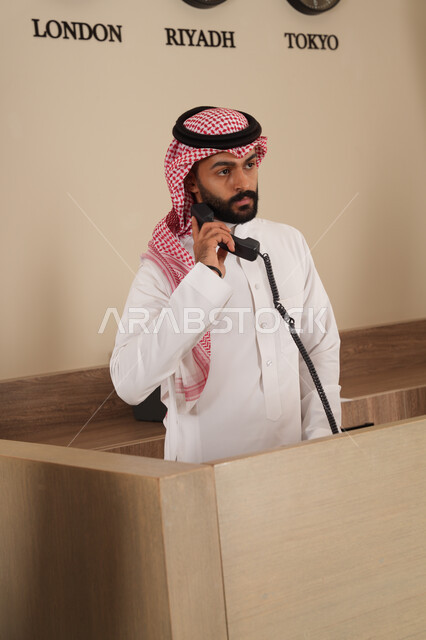 القيام بإجراء مكالمة هاتفية عبر الهاتف, موظف عربي خليجي سعودي يرتدي الثوب والشماغ يعمل في مكتب الاستعلامات, خدمة العملاء في الفنادق الفخمة, اجراءات الحجز والاستفسار, النظر إلى شيء ما والرد على أسئلة الزبائن
