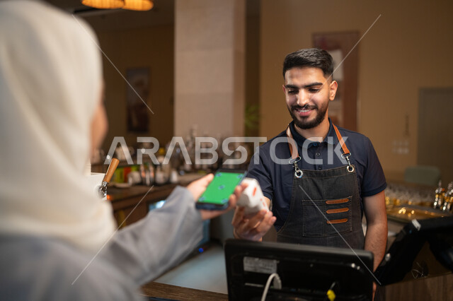 The ease of automatic payment at the cashier, the use of modern technologies in Saudi restaurants, a picture from the back of a Saudi Arabian Gulf woman paying via mobile phone with a blank green screen, chroma, a Saudi young man working in the field of accounting in a cafe