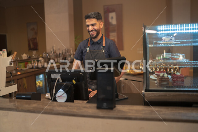 تسجيل طلبات الزبائن على الحاسوب, المقاهي والمطاعم  السعودية, شاب عربي خليجي سعودي مبتسم يرتدي زي العمل يعمل في مجال المحاسبة, بشاشة الوجه والترحيب بالزبائن, الدفع عند الكاشير, استخدام التقنيات الحديثة في الكوفي شوب