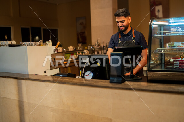 مقاهي ومطاعم المملكة العربية السعودية, الدفع الالكتروني ببطاقة الإئتمان البنكية, شاب عربي خليجي سعودي مبتسم يرتدي زي العمل يعمل في مجال المحاسبة بإيماءات السعادة والسرور, الدفع عند الكاشير, استخدام التقنيات الحديثة في الكوفي شوب