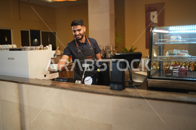 تقديم القهوة للزبون, مقاهي ومطاعم المملكة العربية السعودية, بشاشة الوجه والترحيب بالزبائن, شاب عربي خليجي سعودي مبتسم يرتدي زي العمل يعمل في مجال المحاسبة بإيماءات السعادة والسرور, الدفع عند الكاشير, استخدام التقنيات الحديثة في الكوفي شوب