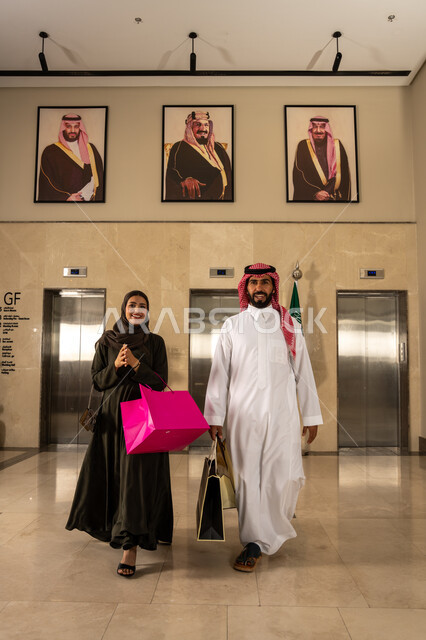 العودة إلى الفندق بعد قضاء وقت ممتع بالتسوق أثناء التخفيضات في اليوم الوطني 23 سبتمبر, رجل وامرأة عربيان سعوديان خليجيان يحملان أكياس التسوق الملونة بإيماءات السعادة والسرور, خلفية صور الملك سلمان والملك عبد العزيز و ولي العهد محمد بن سلمان