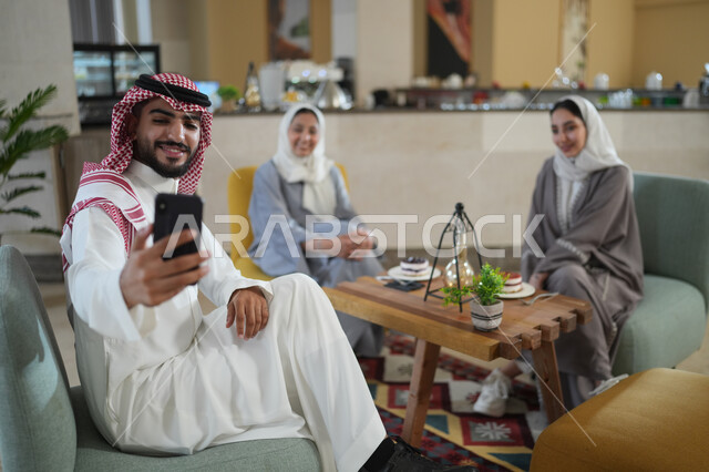 جلسات تعارف وديه, زملاء عمل في اجتماع خارج الشركة, شاب عربي خليجي سعودي يرتدي الشماغ والثوب التقليدي يقوم باستخدام الجوال برفقة أصدقائه, التقاط صور سيلفي تذكارية عبر الجوال, توثيق اللحظات السعيدة, النظر إلى الكاميرا بإيماءات السعادة والسرور