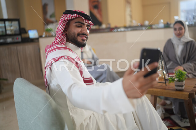 جلسات تعارف وديه, زملاء عمل في اجتماع خارج الشركة, شاب عربي خليجي سعودي يرتدي الشماغ والثوب التقليدي يقوم باستخدام الجوال برفقة أصدقائه, التقاط صور سيلفي تذكارية عبر الجوال, توثيق اللحظات السعيدة, النظر إلى الكاميرا بإيماءات السعادة والسرور