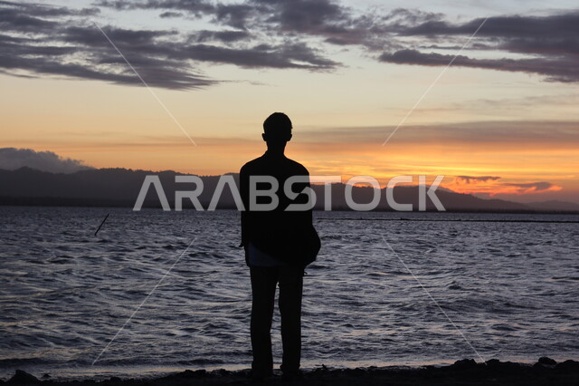 التأمل والنظر في مياه البحر الهادئة، صورة ظلية من الخلف لشاب يقف على شاطئ البحر، الطبيعة البحرية وقت غروب الشمس، أماكن سياحية ترفيهية