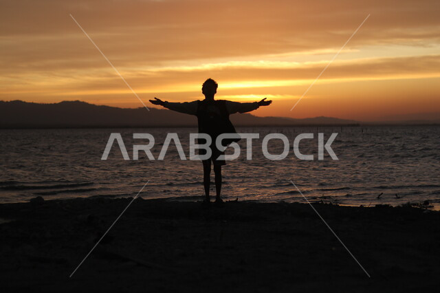 الطبيعة البحرية وقت غروب الشمس، صورة ظلية من الخلف لشاب يقف على شاطئ البحر، التأمل والنظر في مياه البحر الهادئة، أماكن سياحية ترفيهية