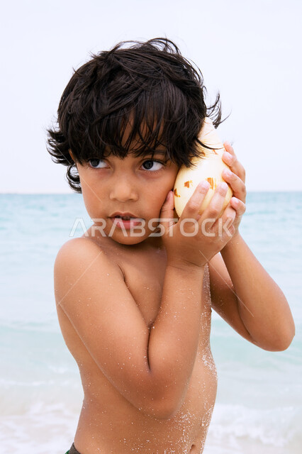  اكتشاف  الصدف والقذائف البحرية، صورة مقربة لولد عربي إماراتي خليجي يقوم بوضع صدفة كبيرة على أذنه ، قضاء أوقات ممتعة ومسلية في العطلة على شاطئ البحر , الشعور بالسعادة والاستمتاع بالفعاليات الصيفية، أماكن سياحية ترفيهية في الخليج العربي.