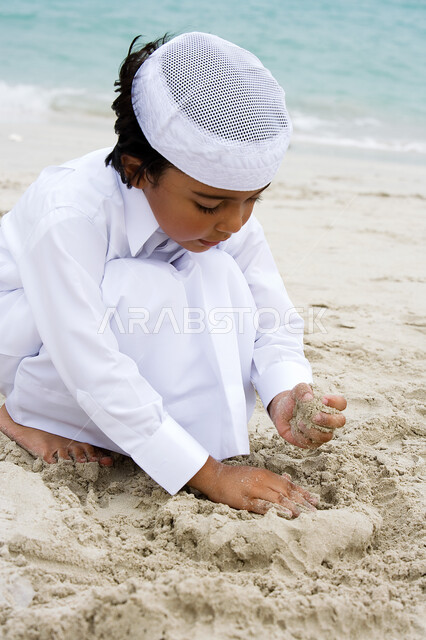 رحلة صيفية ممتعة ، ولد عربي إماراتي خليجي يرتدي الكندورة التقليدية ويلعب بالرمال على شاطئ البحر ،  أنشطه ترفيهية وفعاليات عائلية , إيماءات وجه تدل على السعادة والفرح , منتجعات شاطئية في الإمارات العربية المتحدة .