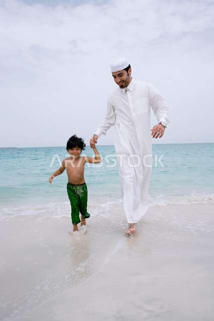 الاستمتاع على شاطئ البحر في إجازه الصيف، أب عربي خليجي إماراتي يرتدي الثوب الاماراتي التقليدي يقوم باللعب والسباحه برفقه ابنه، قضاء عطله ممتعه مع العائله، مشاركه الأباء الأبناء في النشاطات الترفيهية، مفهوم السياحه في دول الخليج العربيه.