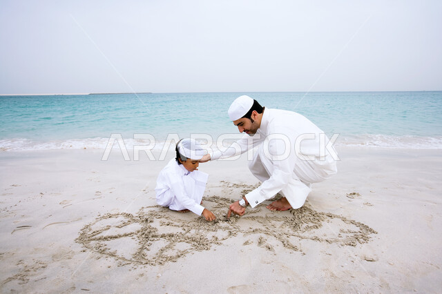 قضاء عطله ممتعه مع العائله، مشاركه الأباء الأبناء في النشاطات الترفيهية، أب عربي خليجي إماراتي يرتدي الثوب الاماراتي التقليدي يقوم بالرسم على الرمال الذهبيه الناعمه برفقه ابنه، شاطئ البحر في إجازه الصيف، مفهوم السياحه في دول الخليج العربيه.