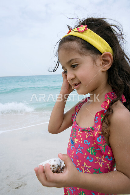 قضاء أوقات ممتعة ومسلية في العطلة على شاطئ البحر ,  صورة مقربة لفتاة عربية إماراتية خليجية ترتدي الزي البحري وتمسك بيدها  صدفة كبيرة  ، اكتشاف  الصدف والقذائف البحرية ،  الشعور بالسعادة والاستمتاع بالفعاليات الصيفية، أماكن سياحية ترفيهية في الخليج العربي.