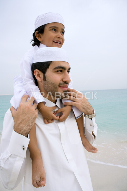 متعه قضاء الإجازه الصيفيه في الساحل، مفهوم الأبوه وشاركه الأبناء نشاطاتهم، التسليه والترفيه بالفعاليات الأسريه الصيفيه، صوره مقربه لأب عربي خليجي اماراتي يرتدي الثوب الاماراتي التقليدي يقوم بحمل ابنه على كتفيه في شاطئ البحر، الرمال الذهبيه الناعمه.