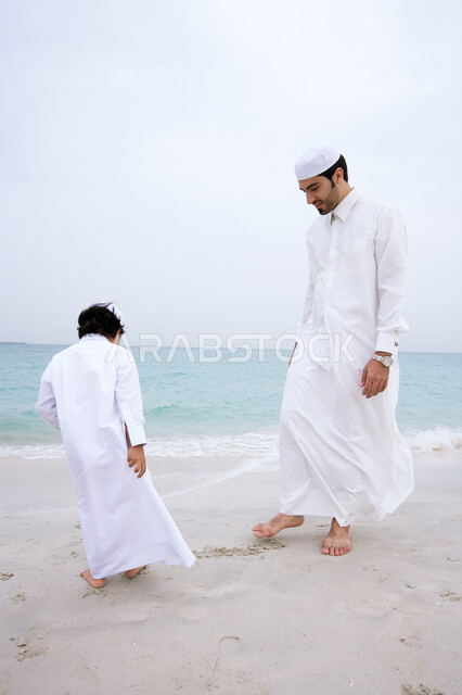 الاستمتاع على الرمال الذهبيه الناعمه، متعه قضاء الإجازه الصيفيه في الساحل، أب عربي خليجي اماراتي يرتدي الثوب الاماراتي التقليدي يقوم باللعب مع ابنه في شاطئ البحر، مفهوم الأبوه وشاركه الأبناء نشاطاتهم، التسليه والترفيه بالفعاليات الأسريه الصيفيه.