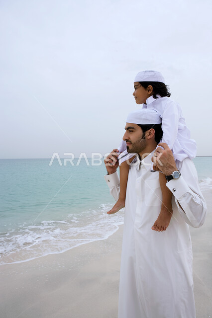 التسليه والترفيه بالفعاليات الأسريه الصيفيه، مفهوم الأبوه ومشاركه الأبناء نشاطاتهم، رجل عربي خليجي اماراتي يرتدي الثوب الاماراتي التقليدي يقوم بحمل ابنه على كتفيه في شاطئ البحر، المشي على الرمال الذهبيه الناعمه، متعه قضاء الإجازه الصيفيه في الساحل.