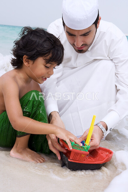 الاستمتاع بالفعاليات الصيفيه، مفهوم السياحه في الشواطئ والمنتجعات الإماراتيه، صوره مقربه لولد عربي خليجي إماراتي يقوم باللعب بالألعاب الرمليه على شاطئ البحر برفقه والده، أجواء عائليه سعيده، قضاء أوقات ممتعه بالتسليه والترفيه مع الأبناء.