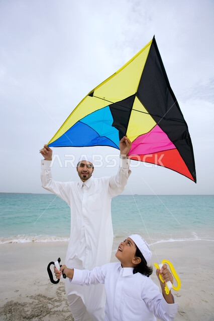 أجواء عائليه سعيده، الاستمتاع بالفعاليات الصيفيه، مفهوم السياحه في الشواطئ والمنتجعات الإماراتيه، ولد عربي خليجي إماراتي يرتدي الثوب الاماراتي التقليدي يقوم بتطير الطائره الورقيه على شاطئ البحر برفقه والده، قضاء أوقات ممتعه بالتسليه والترفيه مع الأبناء.