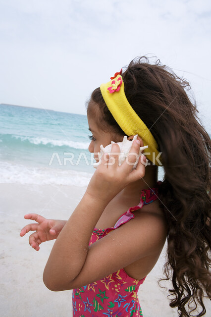 قضاء عطله نهاية أسبوع ممتعه على الشواطئ الرمليه، الاستمتاع بالفعاليات والنشاطات الترفيهية، استكشاف الصدف والمخلوقات البحريه، صوره مقربه لبنت عربيه خليجيه إماراتيه تقوم بوضع قذيفة البحر على أذنها، أماكن سياحيه صيفيه في الخليج العربي.