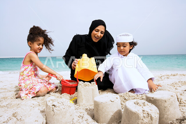 الاماكن السياحيه في الدول العربيه، الأنشطه الترفيهية الصيفية المنوعة والمسليه للأطفال، أم عربيه خليجيه اماراتيه محجبه تقوم بصنع القلاع الرمليه بأدوات الرمل برفقه ابنائها، الاستمتاع باللعب على شاطئ البحر، قضاء عطله ممتعه مع العائله.