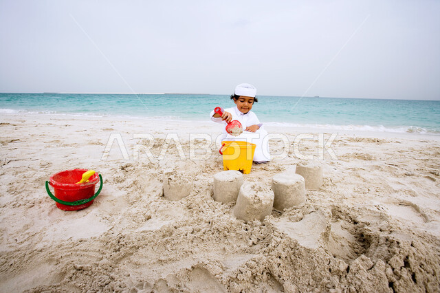 الأنشطه الترفيهية الصيفية المنوعة والمسليه للأطفال، الاستمتاع باللعب على شاطئ البحر، طفل عربي خليجي سعودي يرتدي الثوب الاماراتي التقليدي يقوم بصنع القلاع الرمليه بأدوات الرمل، قضاء العطله في الشواطئ والاماكن السياحيه في الدول العربيه.