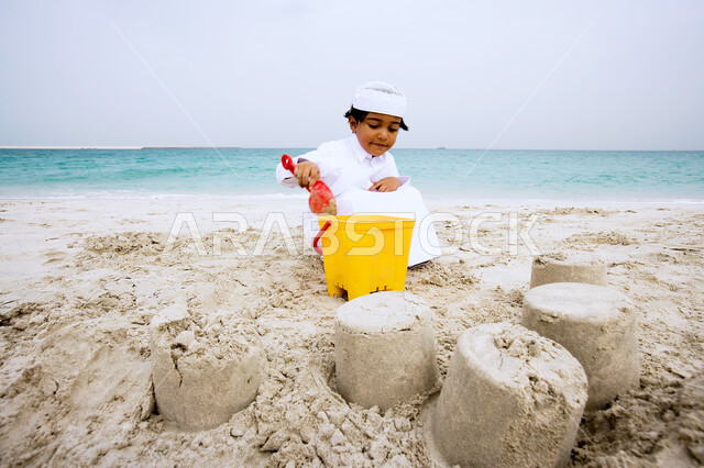 الاستمتاع باللعب على شاطئ البحر، الأنشطه الترفيهية الصيفية المنوعة والمسليه للأطفال، طفل عربي خليجي سعودي يرتدي الثوب الاماراتي التقليدي يقوم بصنع القلاع الرمليه بأدوات الرمل، قضاء العطله في الشواطئ والاماكن السياحيه في الدول العربيه.