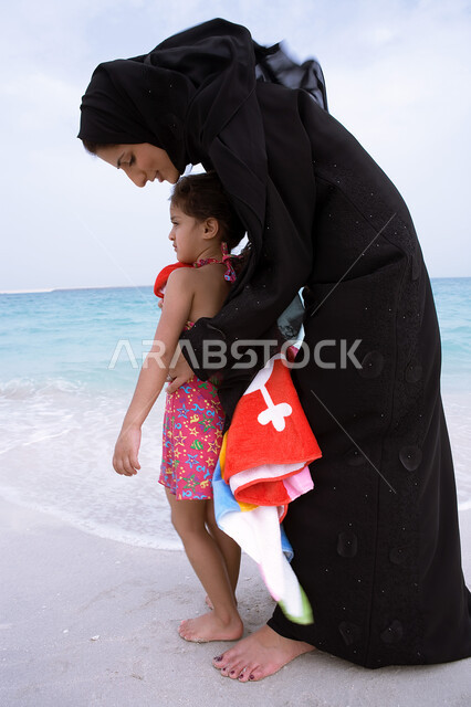 أنشطه ترفيهية عائليه في العطله الصيفيه، إظهار الحب وبناء علاقه صحيه مع الأبناء، مفهوم الرعايه والإهتمام بالأطفال، أم عربيه خليجيه اماراتيه محجبه ترتدي عباءه سوداء تقوم بتجفيف ابنتها بالمنشفة بعد السباحة، أماكن ومنتجعات شاطئيه في الامارات. 