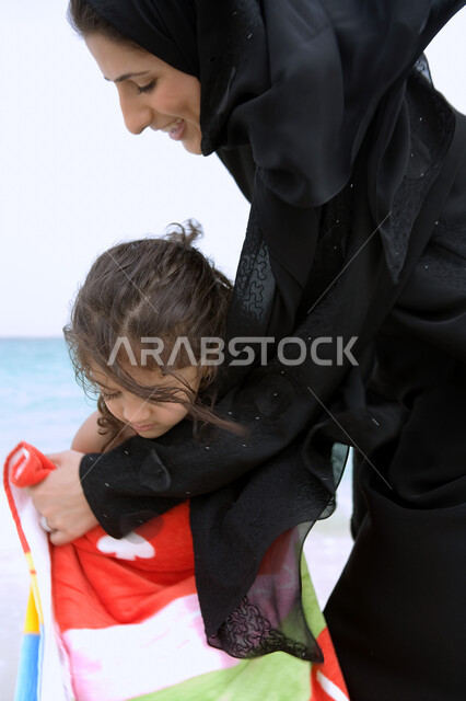 مفهوم الرعايه والإهتمام بالأطفال، إظهار الحب وبناء علاقه صحيه مع الأبناء، أم عربيه خليجيه اماراتيه محجبه ترتدي عباءه سوداء تقوم بتجفيف ابنتها بالمنشفة بعد السباحة، أنشطه ترفيهية عائليه في العطله الصيفيه، أماكن ومنتجعات شاطئيه في الامارات. 