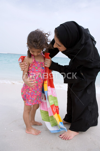 إظهار الحب وبناء علاقه صحيه مع الأبناء، مفهوم الرعايه والإهتمام بالأطفال، أم عربيه خليجيه اماراتيه محجبه ترتدي عباءه سوداء تقوم بتجفيف ابنتها بالمنشفة بعد السباحة، أنشطه ترفيهية عائليه في العطله الصيفيه، أماكن ومنتجعات شاطئيه في الامارات. 
