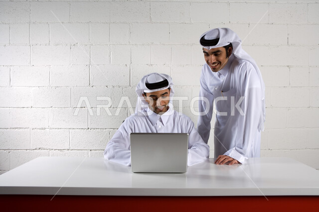 نقاشات وحوارات حول مخططات العمل ، بيئة العمل في الشركات الإماراتية، رجلان أعمال عربيان خليجيان اماراتيان يبتسمان و يرتديان الزي الإماراتي التقليدي ويقومان بدراسة المشروع ، التعاون في تحقيق أهداف الشركة  ، استخدام الحاسوب المحمول , خلفية بيضاء