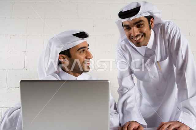بيئة العمل في الشركات الإماراتية، رجلان أعمال عربيان خليجيان اماراتيان يبتسمان و يرتديان الزي الإماراتي التقليدي ويقومان بدراسة المشروع ، التعاون في تحقيق أهداف الشركة ، نقاشات وحوارات حول مخططات العمل ، استخدام الحاسوب المحمول , خلفية بيضاء