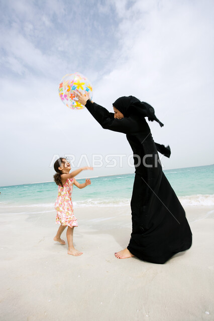 Spending fun summer times in leisure and entertainment with the children, a veiled Arab Gulf Emirati mother wearing a black abaya playing with her daughter with a rubber ball on the seashore, enjoying entertaining activities, the concept of tourism on the coastal beaches in the Emirates.