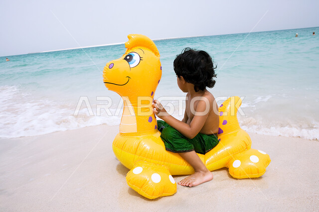 الاستمتاع بالنظر الى شاطئ البحر، الأنشطة الترفيهية والفعاليات الممتعه، قضاء أوقات مسليه في العطله الصيفيه، صبي عربي خليجي اماراتي يجلس على عوامه السباحة، زياره المنتجعات والشواطئ الساحلية السياحية في الدول العربيه.