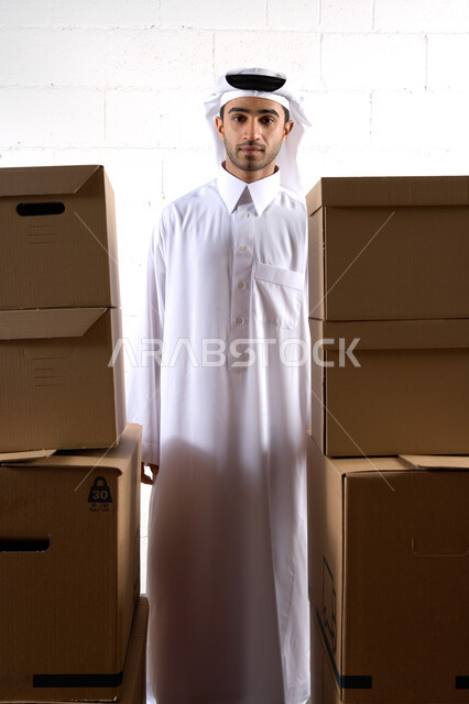 Gestures of satisfaction with fast and safe delivery services, an Arab Gulf Emirati man wearing a traditional dress, arranging cardboard boxes, delivering goods and orders to customers, packing and packaging online shipping cartons, white wall background, full-length body photo