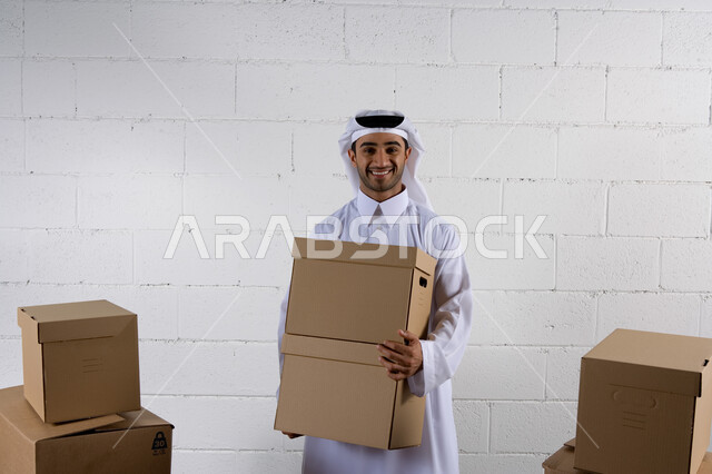 صناديق كرتونية تعبئة وتغليف  ، رجل عربي خليجي إماراتي يرتدي الكندورة التقليدية ويقوم بحمل صناديق الشحن ، كراتين الشحن والتوصيل الأونلاين  ، تسليم البضائع والطلبات للزبائن،  خدمات التوصيل السريع والامن، خلفية رخام ابيض .