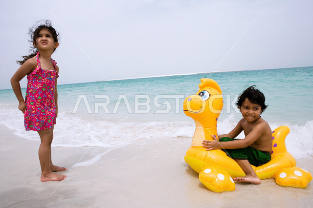 الاستمتاع والتسليه في أوقات العطله، ايماءات الفرح والسعاده للقيام بالسباحة، الأنشطة الترفيهية والفعاليات الممتعه، صبي عربي خليجي اماراتي يجلس على عوامه السباحة في شاطئ البحر برفقه أخته، زياره المنتجعات والشواطئ الساحلية السياحية في الدول العربيه.